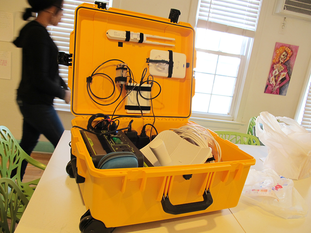 A portable network kit. Yellow hard-shell case containing a router, Raspberry Pi server, antenna, battery, and cables