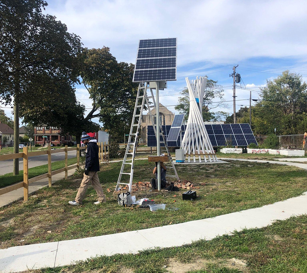 person walking next to a pole with solar panels on the top, with tool bags on the ground below it. More solar panels and a seating area are in the background